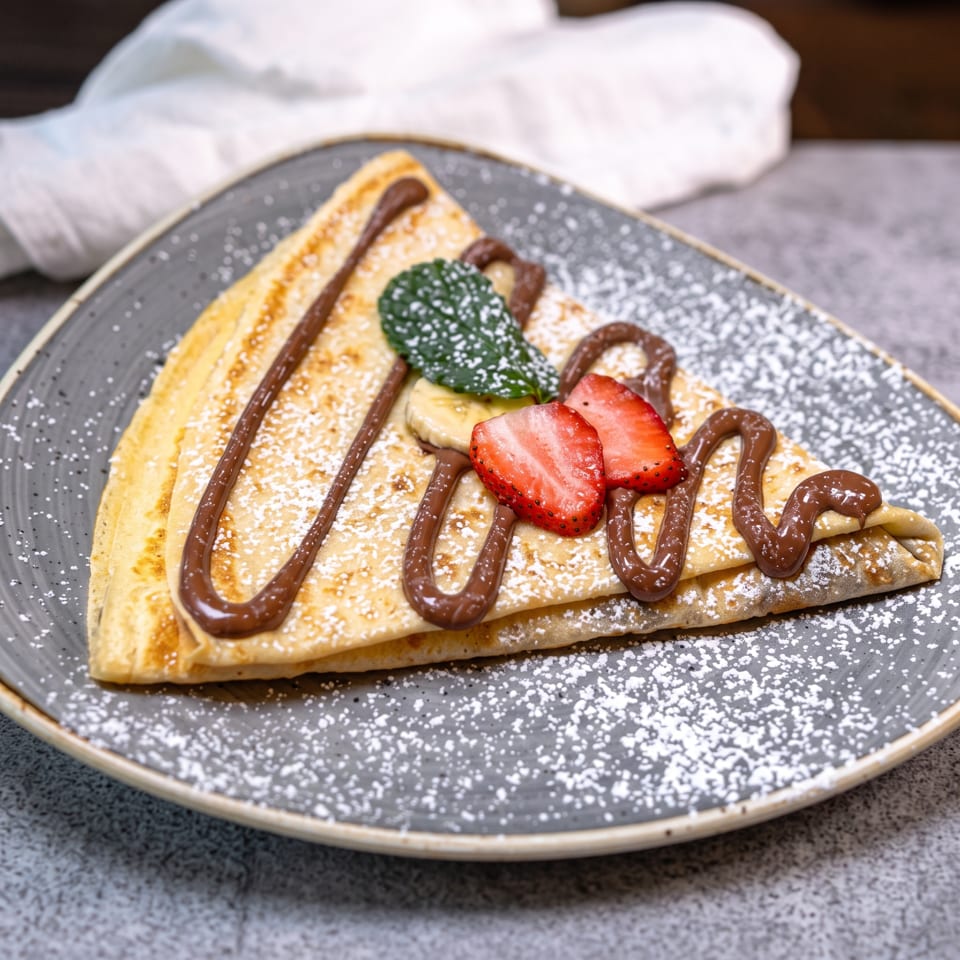 A folded crepe drizzled with chocolate, topped with strawberries, bananas, and mint, dusted with powdered sugar.