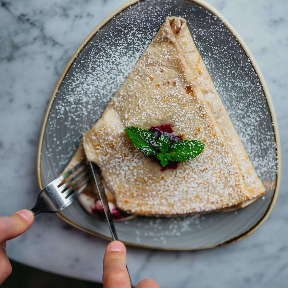 Hands cutting into a powdered sugar-dusted crepe with berry filling and mint on a gray plate.