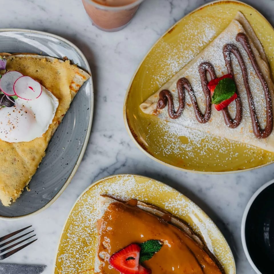 Overhead view of three crepes: one savory with egg, two sweet with chocolate and caramel, and an iced coffee.